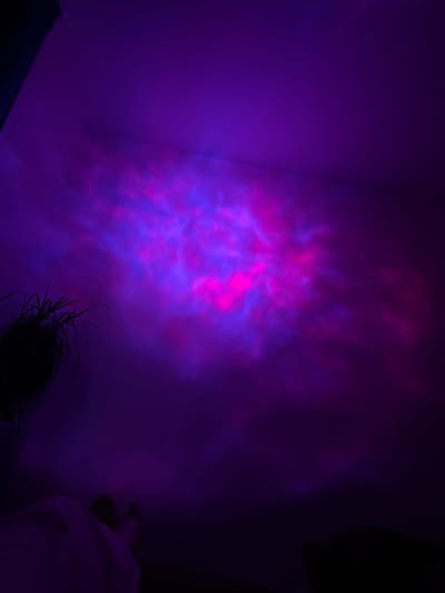 The image depicts a dimly lit room with walls illuminated by a projector displaying swirling patterns of pink and purple light, resembling a nebula or aurora borealis. A dark silhouette of a plant with long, thin leaves is visible in the lower left corner. In the lower portion of the image, a white sheet or blanket is draped over an unseen object, and further to the right, two indistinct figures, likely people, are dimly visible in the shadows. The scene appears to be indoors, with the projector light casting an atmospheric glow. There is no visible indication of the city of Perm, Russia, in this image.