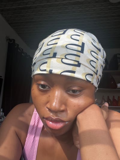 A tanned woman wearing a head wrap with dark blue and light yellow curved shapes posing for a selfie. She is wearing a light pink tank top. Her head is tilted. Her right hand is placed on her cheek. The image is very bright with natural sunlight emphasizing the gloss on her lips and skin. Background elements include window blinds and shelves containing items.