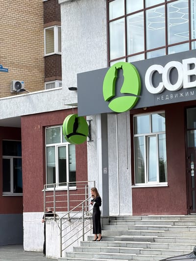 A mid-range shot captures the facade of a building with a burgundy-colored lower section and a lighter-colored, textured upper section. Above a white railing, a woman in a black dress and heels stands on a set of stairs, holding a phone to her ear. To her left, partially obscured by the railing, a man in dark clothing stands near a concrete barrier. A sign with a green owl logo and white lettering, possibly a business name, is visible on the upper part of the building. A smaller, circular green owl sign hangs from a metal bracket on the burgundy section of the building. White-framed windows with blinds are present on both levels of the building. To the left of the building, a portion of a yellow brick structure is visible, with an air conditioning unit mounted on its exterior. A blue street sign with white lettering, reading "УЛ. БОЛЬШЕВИКОВ, 43" (Bolshevikov Street, 43), is affixed to the brick building. The scene appears to be on a street in the city of Tyumen, Russia.