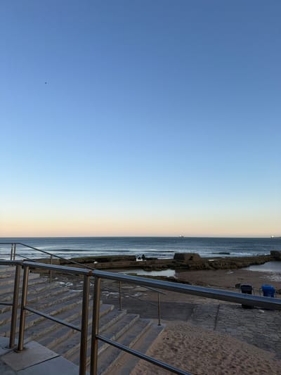 The image depicts a scenic view of the Atlantic Ocean from a paved area with stairs and metal railings, likely a coastal promenade in Cascais, Portugal. The sky is a clear, pale blue, transitioning to a lighter hue near the horizon where it meets the sea. A few small waves are visible in the water, and two cargo ships are present in the distance. The foreground features sand and rock formations along the coastline, with a few concrete structures and two trash receptacles, one blue and one black, to the right. The overall impression is one of a tranquil, coastal setting at either dawn or dusk.