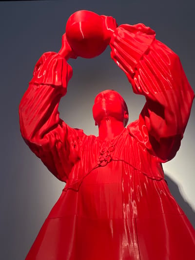 The image depicts a red sculpture of a person wearing traditional clothing and holding a round object in their hands. The sculpture is indoors, positioned against a wall that transitions from a darker shade on the left to a lighter shade on the right. The sculpture's clothing has distinct folds and seams, suggesting it is made of a material that can be molded and shaped. The location is Las Palmas, Spain.