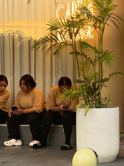 In Kuta Selatan, Indonesia, three individuals are seated on a stepped surface in front of a beige wall. To the left, a person wearing a tan shirt and black pants, with dark hair pulled back, holds a black smartphone. Next to them, another person in a tan shirt and black pants, with dark hair styled in a bun, also holds a black smartphone. To the far right, a third individual, partially obscured and wearing a tan shirt and black pants, is also looking at a smartphone. They are all wearing casual footwear; one has white Crocs, another has black slides with white accents, and the third person's footwear is less visible. In the foreground, a large white planter contains a leafy green plant. A yellow helmet rests on the floor in front of the planter. Behind the individuals, sheer curtains are illuminated by a warm light, and neon lights forming a word, possibly "MASSAGE," are visible in the upper background. The overall scene appears to be an indoor waiting area or lounge.