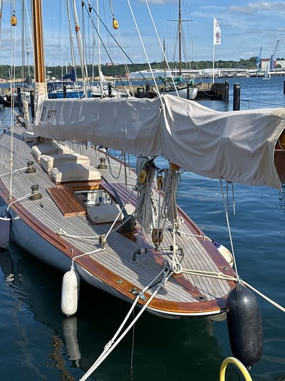 The image captures a close-up, eye-level view of a wooden sailboat docked in the harbor of Kiel, Germany. The sailboat, with its gleaming wooden deck and numerous ropes, is the primary subject. A large, light-colored sail is partially furled and covered with a protective canvas. Several winches are visible on the deck, along with wooden hatches and white fenders tied to the hull. The water is a deep blue, reflecting the bright sky and the sailboat. In the background, other boats and industrial structures, including cranes and buildings, suggest a bustling port environment. The overall scene is one of a well-maintained vessel at rest in a maritime setting.