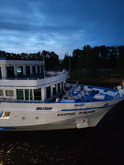 The image shows a side view of a white and blue passenger ship named "АНДРЕЙ РУБЛЁВ" (Andrei Rublev) on dark water at dusk. The ship has multiple decks with windows and railings, and lights are visible on the upper decks. Several people are present on the decks of the ship. The background features a dark line of trees and a deep blue sky with clouds. This scene is likely taking place on the Sheksna River, which flows through the city of Sheksna in Russia, given the presence of the river and the name of the ship which is associated with Russian history and culture. The lighting suggests it is evening or early morning.