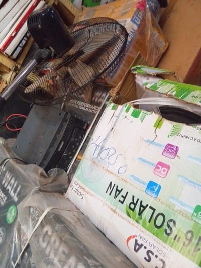 The scene depicts a cluttered indoor storage or retail area in Ankpa, Nigeria, filled with electronic goods and their packaging. In the upper left, a partially assembled black standing fan with a metal cage is visible. Directly below it, a black electronic inverter or amplifier with red wiring is situated. The foreground is dominated by stacked cardboard boxes; a prominent one on the right is labeled "C.S.A 16" SOLAR FAN," featuring graphics for "DC INPUT," "AC INPUT," and "Fan Function," with handwritten blue script resembling "K800" on its surface. Other boxes, some wrapped in clear plastic, display labels such as "HIGH PERFORMANCE SOLAR TALL TUBULAR BATTERY" and "WALL," indicating merchandise primarily related to solar energy and batteries. No individuals are present, and no actions are observed.