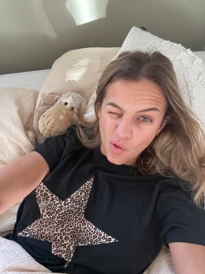 A female subject is depicted lying in bed in a room within Veauchette, France. She has light brown hair, is wearing a black t-shirt with a leopard-print star design on the chest, and small hoop earrings. Her right eye is closed in a wink, and her lips are puckered. A light-colored stuffed animal, resembling a sheep, lies next to her left shoulder on a light beige pillow. Additional pillows, one with a textured pattern, and a light-colored duvet are visible. A light gray wall forms the background, exhibiting a bright rectangular reflection consistent with natural light from an unseen window.
