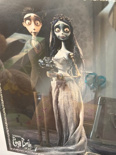 The image displays two stylized figures, reminiscent of characters from stop-motion animation, positioned in a somewhat eerie, gothic setting. The figure on the left appears to be a man with wide, startled eyes, dark spiky hair, and wearing a dark suit with a white cravat. The figure on the right is a woman with pale blue skin, large, dark-rimmed eyes, black hair cascading down, and wearing a tattered, white wedding dress adorned with delicate lace. She has a flower bouquet clutched in her hands. The background is dark and appears to be a room with textured wallpaper and some indistinct objects. The overall impression is that of a display or a promotional image for a film or product, likely related to "Tim Burton's Corpse Bride," as evidenced by the partially visible text at the bottom left of the image. There is no discernible location in Alicante, Spain, or any other specific geographic location visible in the image.