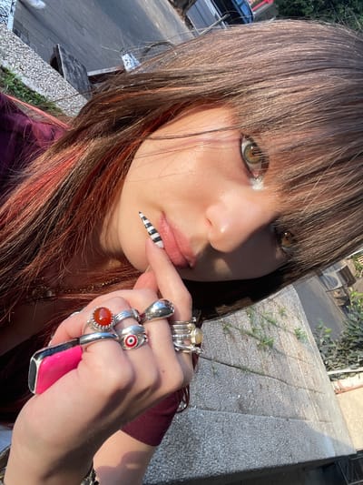 A medium shot captures a young woman with long, brown-auburn hair styled with blunt bangs. She has fair skin, hazel eyes accentuated with makeup, and is adorned with numerous rings on her fingers, including one with a pink stone and others with decorative stones. Her left hand is raised to her lips, with a black and white striped false nail touching her lower lip. The background suggests an urban setting, with a low concrete wall and a glimpse of a tiled sidewalk or patio. The scene appears to be outdoors in daylight, possibly in a residential or commercial area in Raanana, Israel. A bench is partially visible in the background.