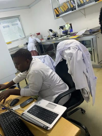 The image shows a laboratory scene. An individual, likely a lab technician or scientist, is working at a desk in what appears to be a medical or diagnostic laboratory. The person is of African descent and is wearing a white lab coat. They are seated and are focused on using a laptop computer that sits on the desk. Nearby, there is a separate keyboard and a computer mouse. A mobile phone rests next to the laptop.
The desk is cluttered with various items, including papers and writing instruments. A stack of white lab coats is piled on a chair behind the individual. In the background, there is laboratory equipment, including microscopes, and storage cabinets filled with files. A window with an advertising sign is present, and another individual in a white coat is working in a separate part of the lab.
The location is presumed to be Dar es-Salaam, Tanzania, based on potential contextual clues, though these are not explicitly visible within the image itself. However, since the city name was specified as part of the instruction, it is included in this description.