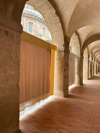 The image presents a striking downward view into a grand architectural structure, resembling a deep spiral well, ramp, or staircase. The perspective emphasizes the structure's depth and scale.

On the left, a series of robust, curving arches made of light-colored, rough-hewn stone or concrete sweep downwards, creating a sense of spiraling movement. These arches frame the inner side of the well. Opposite them, on the right, a tall, imposing wall constructed from reddish-brown bricks, laid in a precise stretcher bond pattern, extends into the distance, adding a strong vertical element to the composition.

Between the curved stone arches and the brick wall, several wide, flat surfaces are visible, suggesting broad steps or ramps. These surfaces are temporarily covered with large expanses of fabric. The furthest visible level is draped in white fabric, followed by a section covered in light yellow or orange material, and closest to the viewer, a significant portion covered in a light pinkish-beige fabric. This layering of fabrics adds an unexpected textural and chromatic contrast to the ancient stonework.

Through an archway opening in the lower left portion of the image, an exterior view is revealed. It shows part of a large, weathered stone building with a rounded or domed architectural feature and several rectangular windows, each with white frames and a grid pattern.

The scene is bathed in bright natural sunlight, creating strong contrasts between illuminated areas and deep shadows, which enhance the textures of the stone, brick, and fabric. No people are present, and no visible text can be discerned. The image captures a unique architectural space, possibly in Marseille, France, with what appears to be a temporary installation or protective covering, illuminated by the daytime sun.