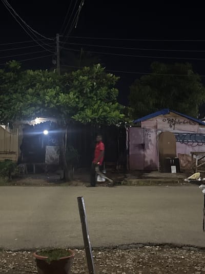 In Abuja, Nigeria, at night, a street scene unfolds with a focus on two small structures. The structure on the left appears to be a kiosk or shop with a corrugated metal roof, partially obscured by a large tree with lush green foliage. A bright light shines from under the roof of this structure. To the right, a second structure, also with a corrugated roof, is painted pink and features dark blue trim. This building has several markings, including what appears to be a phone number "080646069" and another set of numbers and letters, "703069361". A person wearing a red shirt and dark pants is walking across the asphalt road in front of these structures. The road is lined with gravel on the sides. In the foreground, a potted plant sits on the gravel, next to a dark vertical pole. Electrical wires are visible crisscrossing the dark night sky above the scene.