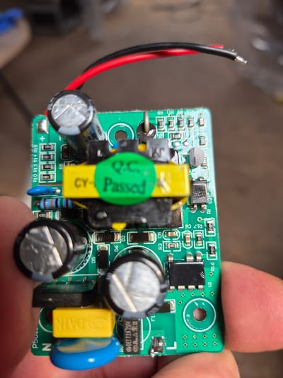 The image shows a close-up, slightly blurry view of a green printed circuit board (PCB) with various electronic components. The PCB is held in the palm of a person's hand. Prominent components include several electrolytic capacitors (one with a silver top and a cross-shaped indentation, two larger ones with black casings and silver tops), a yellow film capacitor with "CY- Passed" printed on it in green, a black chip with multiple pins, resistors, and surface-mount components. Red and black wires are connected to the top of the PCB. The background is out of focus and appears to be an indoor setting with a blurred wooden surface. There is no visual information in the image that relates to the city of Masów, Poland.