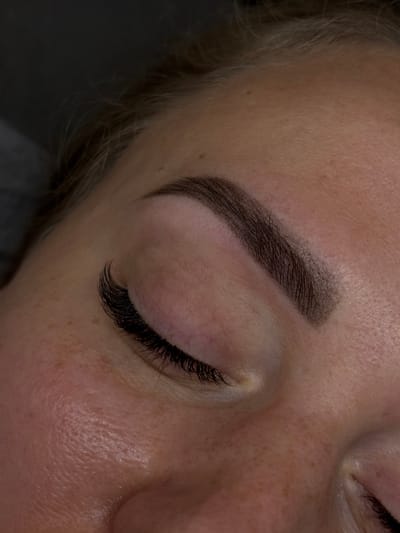 A close-up, high-angle, eye-level shot shows a person's closed eye with a perfectly shaped eyebrow and voluminous eyelashes. The skin is fair with some freckles visible. The overall lighting is soft, and the background is dark and out of focus, suggesting an indoor setting. The image does not contain any information to determine the location in the city of Khimki, Russia.