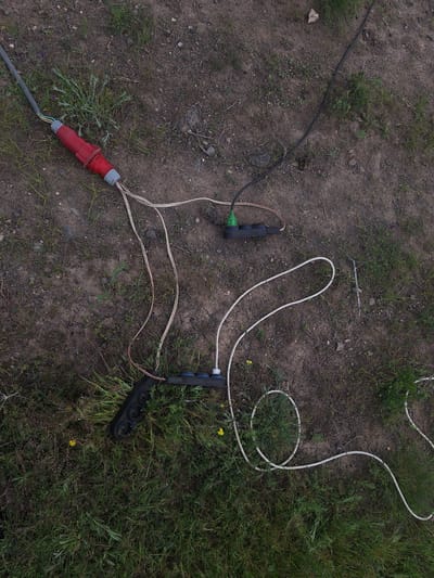 The scene depicts an unidentifiable location with ground covered by a mix of dirt and grass. Several electrical components are scattered across the area. A large, red industrial plug is connected to a gray cable, extending towards the lower-left corner. Two black power strips are visible, each with three sockets. One strip is connected to a green plug, while the other is linked to a white cable that loops around in a coil. No people are present in the frame. The location in Tlik, Armenia is not identifiable from the given image.