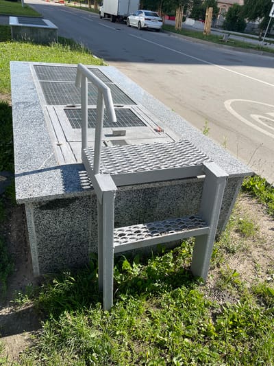 The image shows a concrete structure with a granite-like finish, covered by metal grates and featuring a metal ladder with textured steps. The structure is situated outdoors, adjacent to a paved road and surrounded by grass and dirt. In the background, a white truck and a white sedan are visible on the road, along with trees and buildings. The environment appears to be urban. A speed limit sign is partially visible on the right side of the road. There are no people visible in the image. This scene is likely located in a public area in the city of Zakharino, Russia, possibly near infrastructure related to utilities or drainage.
