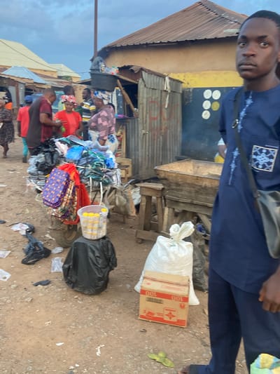 In Bukuru, Nigeria, a street market scene unfolds. A young man in a blue garment stands in the foreground, his gaze directed forward. Behind him, a cart is laden with various items, including bags and what appear to be packaged goods. Several other individuals are present, some engaged in commerce or interacting with the goods on display. The environment suggests a bustling marketplace with makeshift stalls and a dusty, outdoor setting. The sky above is overcast.