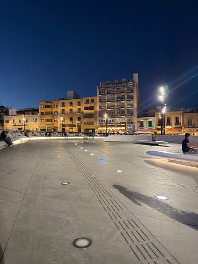 This is an eye-level, vertical shot of a public square at dusk in Nicosia, Cyprus. The scene is dominated by a large, paved area with embedded lighting that casts a warm glow on the textured concrete. A prominent tactile paving strip runs diagonally across the pavement, indicating a path for visually impaired individuals.

In the background, several multi-story buildings line the square. The buildings are constructed with light-colored stone and feature numerous windows, suggesting residential or commercial use. Some of the windows are illuminated, hinting that people are inside. Streetlights with multiple bulbs are visible, adding to the ambient lighting of the scene.

Several people are present in the square, scattered across the paved area and near the buildings. Some are sitting, while others appear to be walking or standing. One person in the upper right corner is captured in mid-action, possibly taking a photo with their phone. The mood of the scene is relaxed and calm, suggesting a leisurely evening in a public space.

The time of day is dusk, as indicated by the dark blue sky and the artificial lighting illuminating the square. There are no obvious weather cues, but the lighting suggests clear conditions. No specific event is apparent; it appears to be a typical evening in a city square.

A sign with the name "GIOZOUDI" is partially visible on one of the buildings in the lower left quadrant of the image, providing a potential location cue. The overall impression is one of urban life at twilight, with the interplay of natural and artificial light creating a serene atmosphere.