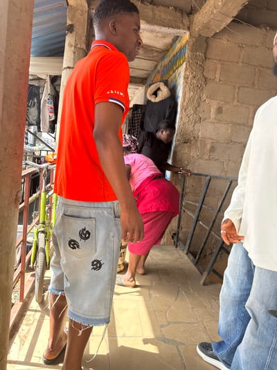 In Bukuru, Nigeria, a man in an orange polo shirt and distressed denim shorts stands on a paved walkway. To his right, another man in a white shirt and blue jeans is partially visible. Behind the first man, a woman in a black shirt and pink skirt is seen entering a doorway with a fur-lined coat hanging inside. Another woman, wearing a pink headscarf and pink top, is positioned in front of the doorway. A green bicycle is propped against a railing to the left. The environment appears to be an outdoor market or residential area, with exposed brick walls, corrugated metal roofing, and various items hanging on display in the background. The sunlight casts shadows on the ground.