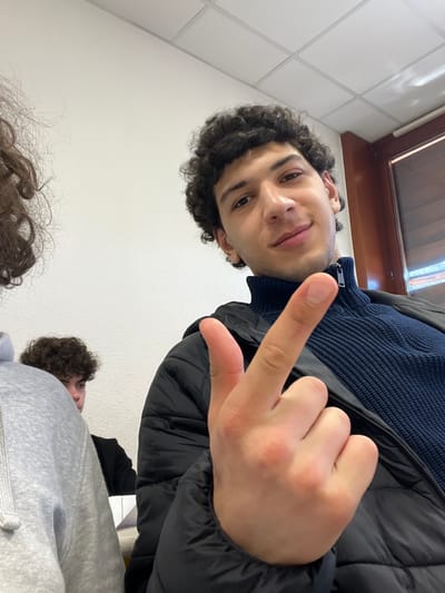 A selfie-style image of a young man with dark curly hair, wearing a navy blue turtleneck sweater and a black puffer jacket. He is holding up his middle finger. Two other individuals with curly hair are partially visible at the bottom of the frame. The background suggests an indoor setting, possibly a classroom, with white tiled walls and a window in the upper portion of the image. The location in Schiltigheim, France, is not discernible from the visual data.