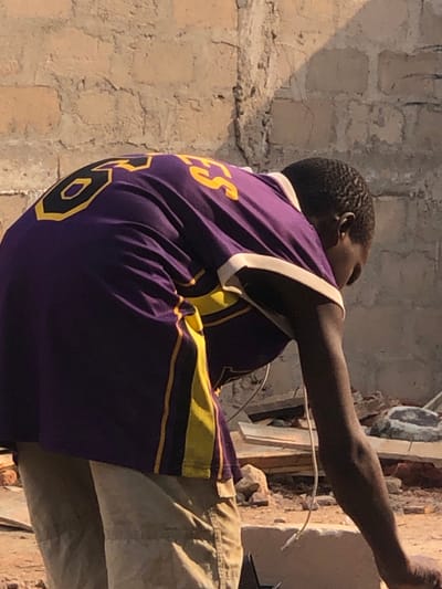 A male individual with dark skin and short dark hair is depicted bending forward and looking downwards, engaged in an unspecified activity. He is wearing a purple short-sleeved shirt with yellow and black numbers "8" and letters "ES" visible on the back, along with yellow vertical stripes on the sides and white trim on the collar and sleeve ends. A white cable, consistent with a headphone or charging cord, hangs from his right side near the ear area. He wears tan-colored pants or shorts. The scene is outdoors, set against a background of a light-colored, weathered concrete block wall showing visible mortar lines and some cracking. The ground beneath him is uneven, dusty, and strewn with construction debris, including rocks, pieces of wood, and fragmented materials. The lighting suggests strong sunlight from an overhead source, casting shadows on the wall and the ground. This scene takes place in Enugwu-Ukwu, Nigeria.