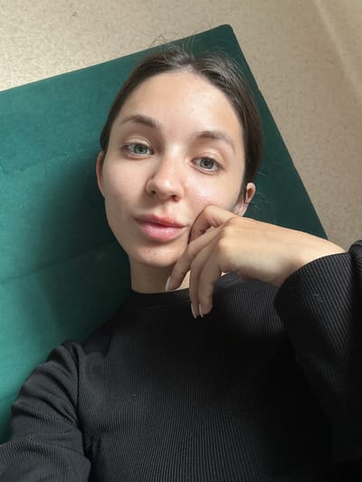 This media file features a close-up, overhead shot of a young woman lying down. She is wearing a black, ribbed long-sleeved shirt. Her face is turned towards the camera, and her left hand is gently touching her cheek. Her eyes are open and looking forward, and her lips are slightly parted. The woman has fair skin with some light freckles, and her eyebrows are dark. Her hair is dark brown and pulled back. She is lying on a dark green mat, which is situated on a light-colored, textured floor. The lighting suggests it might be daytime, with natural light illuminating her face. The overall mood appears relaxed and contemplative. There is no visible text in the image. The location context provided is Sverdlovskiy, Russia.