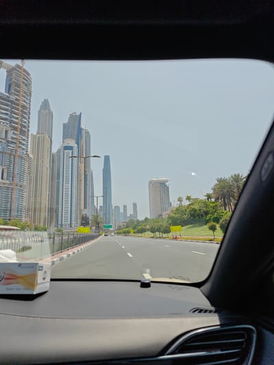 The image, taken from inside a vehicle, shows a view of Dubai Marina. Tall skyscrapers dominate the skyline on the left, with several under construction. A multi-lane road stretches ahead, bordered by a metal fence and a green park area with palm trees on the right. Traffic lights and street signs are visible along the road. The sky is clear and bright, suggesting a sunny day. In the foreground, a box of tissues rests on the car's dashboard. The overall scene depicts a modern urban landscape in Dubai.