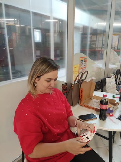 A woman with blonde hair is sitting at a table in what appears to be a casual dining or entertainment establishment in Navapolatsk, Belarus. She is wearing a red top with small white polka dots and is opening a white box with red lettering that reads "Raffaello". On the table in front of her are an open pizza box, a bottle of dark soda, a mobile phone, and other miscellaneous items. The environment suggests an indoor space with large windows and modern interior design, possibly a cafe or play center.