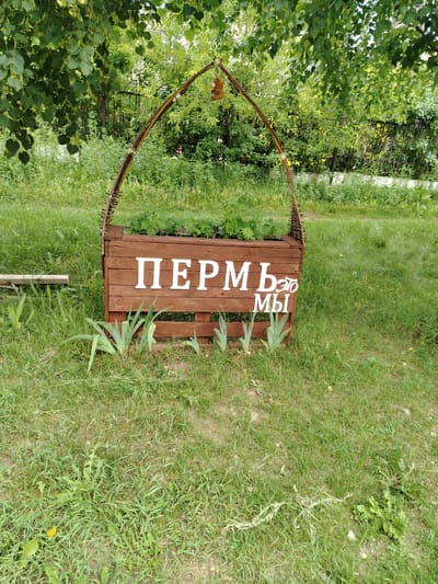 This image captures a wooden planter box decorated with white lettering that reads "ПЕРМЬ это МЫ" (Perm is Us). The planter box is situated outdoors in a grassy area, possibly a park or garden, with lush green foliage in the background, including trees and a fence. A decorative arch, made of natural materials and adorned with a small, brown stuffed animal, is attached to the planter. The scene appears to be a public space, possibly in the city of Perm, Russia, given the inscription. There are no people visible in the image.