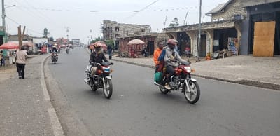 This image captures a street scene in Goma, Democratic Republic of the Congo. The scene is dominated by a paved, dark grey asphalt road, appearing worn.

In the mid-ground, two motorcycles are prominent, both moving towards the viewer. The motorcycle on the left is silver and maroon, operated by a male driver wearing a dark top, light trousers, and a patterned headwrap. The red motorcycle on the right carries a male driver in a grey hoodie and pink helmet, transporting two passengers; the first is a male in an orange hoodie, and the second, partially obscured, wears red/brown clothing. A green bag is affixed to the side of the red motorcycle.

On the left sidewalk, a male pedestrian walks away from the foreground, balancing three long wooden poles vertically on his head.

The street is bordered by single and multi-story concrete and brick buildings. On the right side, structures include a building with a large wooden board leaning against its facade and another under construction, featuring exposed concrete frames. Several small shops and market stalls with red umbrellas are visible along both sides of the road. Scattered pedestrians and additional motorcycles are present further down the street, indicating a busy thoroughfare. Overhead power lines and utility poles are visible against an overcast sky.