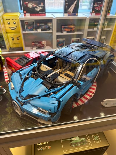 The image displays a collection of LEGO models in what appears to be a retail display case. The primary subject is a large, partially assembled blue LEGO supercar, resembling a Bugatti Chiron, with its intricate mechanical details exposed. Next to it, a red LEGO sports car is visible. In the background, shelves contain various other LEGO set boxes, including a white and red Formula 1 car, a rocket, and an orange sports car. The display case is illuminated, reflecting on the surfaces of the LEGO models and the glass. The lower portion of the image shows a box with a detailed illustration of a green military tank, labeled "1:1" and "26.6CM". The context suggests this is a toy store or a hobby shop. There is no information in the image to indicate a specific location within Yekaterinburg, Russia.