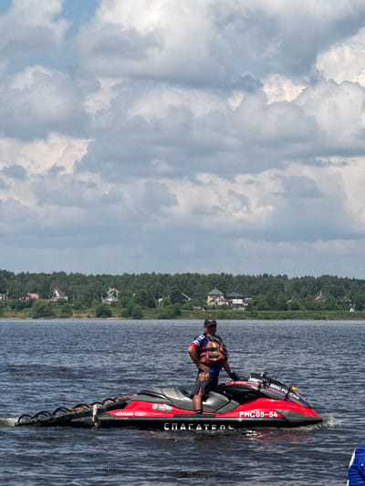 A man in a life vest stands on a red jet ski in the water. The jet ski has Russian writing on it. In the background is a tree line and some houses. The sky is cloudy.