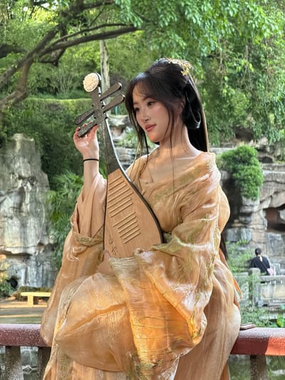 In an outdoor garden setting, likely in Yuzhong District, China, a young woman is seated on a railing, holding a traditional Chinese lute. She is dressed in an elaborate, flowing peach-colored gown with golden embroidery. Her dark hair is styled with ornate gold ornaments. The lute, made of wood, has four strings and a decorative white embellishment at the top. The background features lush green foliage, rock formations, and water, suggesting a traditional Chinese garden or park. Sunlight filters through the trees, illuminating the scene. In the distance, a person in a black shirt is visible walking up stone steps. The overall impression is one of serene beauty and cultural representation.