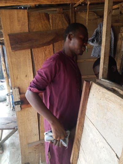 This image depicts an indoor or semi-sheltered area constructed primarily from various wooden planks and beams in Adankolo, Nigeria. The central figure is a dark-skinned male, likely an adult, positioned in profile facing right. He wears a maroon long-sleeved tunic, with the right sleeve rolled up to just below the elbow. His gaze is directed forward or slightly downward. In his right hand, he holds a small, rectangular object wrapped in transparent plastic, possibly a bundle of items. To the upper right background, a patterned, light-colored fabric or garment hangs on a wooden beam. The partially visible head of a second, dark-skinned individual is discernible in the lower right background, behind a light-colored wooden structure, looking downwards. The environment appears to be a workshop or storage area, with varying shades and textures of wood forming walls and structural elements. A metal latch is visible on a wooden panel in the left mid-ground. Natural light illuminates the scene, creating soft shadows.