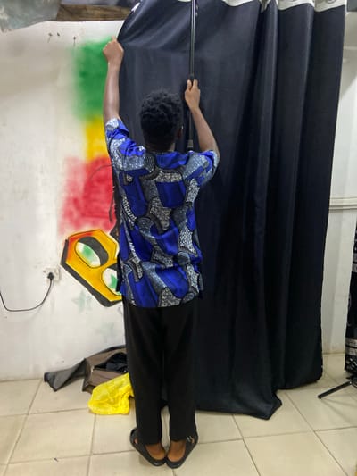 The image captures an indoor scene in Mararaba, Nigeria, featuring a person viewed from the back, manipulating a black fabric curtain. The individual, dressed in a blue, black, and white patterned short-sleeved top and black trousers with black slides, has dark skin and short dark curly hair. Their arms are raised, with the left hand gripping the top edge of the black curtain, pulling it to the left, and the right hand holding a black metal pole that supports the curtain.

Behind the curtain, on the left side of a light-colored wall, a graffiti-style artwork is visible. This artwork includes vertical bands of green, yellow, and red paint, beneath which is a yellow and black geometric design resembling stylized eyes with green pupils. A white electrical outlet with a single socket is mounted on the wall below the artwork, with a black cable extending downwards from it.

The floor is covered with light-colored square tiles with dark grout. On the lower left, an open dark-colored bag rests on the floor next to a crumpled bright yellow plastic bag. The black curtain, featuring a white/light grey trim at the top, obscures the right portion of the wall. A black metal stand leg is partially visible on the far right of the tiled floor, suggesting a support structure for the curtain or other equipment.