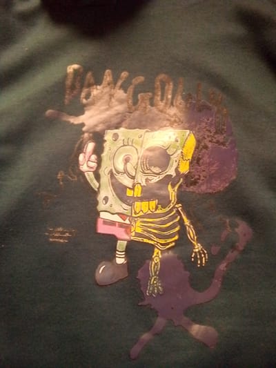 The image is a close-up shot of a dark green t-shirt with a graphic print. The print features a stylized Spongebob Squarepants character that is half living and half skeletal. The living side of Spongebob is green with his signature eyes, nose, and mouth. The skeletal side reveals a ribcage and arm bones in yellow and white. The character is depicted with a raised hand, seemingly giving a middle finger. Around the character, there are splotches of purple and white paint, suggesting a grunge or artistic design. The text above Spongebob appears to be in a distressed, gold-colored font, but it is largely obscured by the white paint splotches, making it illegible. The t-shirt is made of a textured fabric, and the print shows signs of wear or a deliberate distressed aesthetic. The location of Barakin Akawo, Nigeria is not discernible from the image.