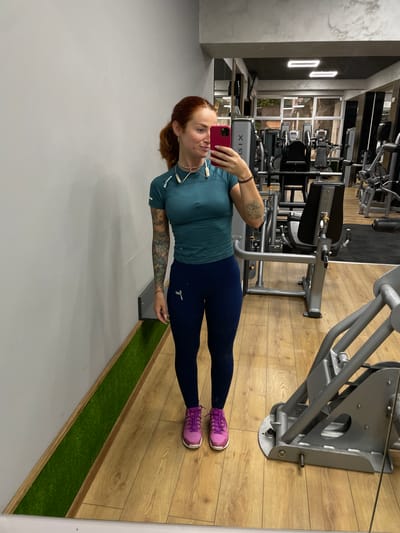 A woman with red hair pulled into a ponytail stands in a gym, taking a selfie in a mirror. She is wearing a teal athletic top and dark blue leggings, with bright purple athletic shoes. Her arms are covered in tattoos. She has headphones around her neck and is holding a pink smartphone. The gym has wooden floors, mirrors, and various weight machines visible in the background. A strip of artificial grass runs along the base of the wall to her left. The gym appears to be located in Pernik, Bulgaria, as indicated by the watermark on the image.