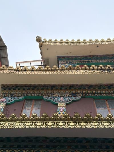 The image shows an upward view of a building's exterior, likely a temple or monastery, with intricate golden decorations and colorful architectural elements. The building features layered roofs with ornate golden trim, resembling floral motifs and repeating patterns. Below the golden trim, there are sections with detailed paintings and carvings in vibrant blues, greens, reds, and yellows. Two windows are visible, set within a dark brown facade. The sky above is overcast and light gray. The architectural style suggests a Tibetan or Buddhist influence, consistent with religious structures found in Dharamshala, India, a city known for its significant Tibetan population and monasteries.