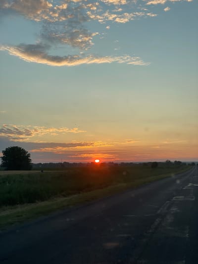 The image shows a rural landscape at sunset, likely in Onițcani, Moldova. A paved road curves into the distance on the right side of the frame. To the left of the road, there is a field of grass and some trees. The sun is setting on the horizon, casting a warm glow over the scene. The sky is a gradient of orange, pink, and blue, with scattered clouds. There are no people or animals visible in the image. The overall impression is one of tranquility and natural beauty.