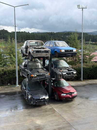 This image displays a vehicle dismantling facility in Vila Real, Portugal, showcasing six severely damaged and partially disassembled vehicles arranged on a three-level metal racking system on a wet concrete surface.
On the left stack, from top to bottom: A light grey/white BMW 2002 coupe, stripped of its doors, hood, and interior, exhibiting extensive rust. Below it is a black Ford Escort Mk4 hatchback, missing its front bumper, with a damaged hood and an open sunroof. The lowest vehicle is a dark grey/black Alfa Romeo 156 Sportwagon with severe front-end impact damage, lacking its bumper and headlights, and a crumpled hood.
On the right stack, from top to bottom: A blue Talbot Horizon sedan, elevated on supports and missing its wheels. Beneath it is a dark grey/black sedan, with its hood removed, exposing the engine bay, and a missing front bumper. The bottom vehicle is a red Alfa Romeo 156 sedan, presenting substantial front-end collision damage, a crumpled hood, and a compromised driver's side front wheel.
The immediate ground is wet, grey concrete or asphalt. The background features a dense evergreen forest covering a hillside under a heavily overcast, cloudy sky. Two tall metal light poles with floodlight fixtures are visible, flanking the car rack. Additional salvage vehicles are discernible in the distant background.