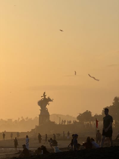 A silhouetted scene unfolds at sunset on a beach in North Kuta, Indonesia. A large, elaborate dragon-like statue, perched atop a multi-tiered structure, dominates the mid-ground, silhouetted against the hazy, orange sky. Numerous people are scattered across the sandy beach, some walking, others sitting or interacting in small groups. In the distance, a line of trees and buildings create a hazy backdrop. Two long, ribbon-like kites are visible in the sky, along with a few small birds. The overall impression is one of a tranquil evening gathering on a tropical shore.