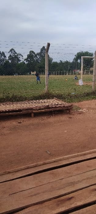 The image shows a scene in Lwandeti, Kenya, located at latitude 0.563 and longitude 34.806. The scene is a dirt field with a wooden structure in the foreground. There is a wooden bench in the middle ground. In the background, there is a green field, a fence, and trees. Several people are visible in the distance. The people are walking and playing in the field. The scene is taken from a slightly elevated vantage point, looking towards the field. The weather is cloudy.