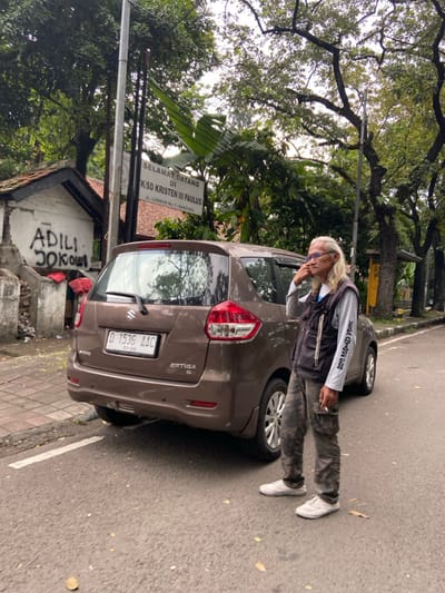 The image shows a man standing next to a brown Suzuki Ertiga car parked on a paved street in front of a building in Bandung, Indonesia, located at latitude: -6.911, longitude: 107.618. The man is wearing a grey t-shirt with black writing on it, a black vest with silver chains hanging from the zipper, and camo cargo pants. He is holding a cigarette in his right hand and looking to the side. The car has a license plate that says "D 1536 AAC". The building in the background has a sign that reads "Selamat Datang Di KSD Kristen III Paulus JL. Lombok No. 7 - Bandung". There is graffiti on the wall of the building that reads "ADILI DOKOL". The street is paved and has a white line marking the edge of the road. There are trees and plants in the background. The sky is overcast.