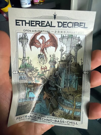 The image shows a poster for the "ETHEREAL DECIBEL" OPEN AIR FESTIVAL, which is scheduled to take place from July 2nd to 5th, 2026. The poster features an intricate illustration of a futuristic, possibly alien, cityscape with towering structures, lush vegetation, and a central winged creature. The festival genres listed are PSYTRANCE, TECHNO, BASS, and CHILL. The bottom of the poster indicates the location as "LE PARC - SAINT SYMPHORIEN DES MONTS (50)". The poster is held by a person's hand, with a portion of the hand visible in the foreground. The background is blurred and appears to be indoors, with some objects on a shelf. The question asks to include the location in the city of Lyon, France, however, the poster only states "LE PARC - SAINT SYMPHORIEN DES MONTS (50)". Saint Symphorien des Monts is a commune in the Manche department in northwestern France, and there is no indication that this location is within the city of Lyon.