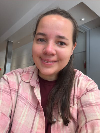 A front-facing, waist-up selfie depicts a young adult female smiling in an indoor setting identified as being in the city of Osaka, Japan. The subject has fair skin, dark brown hair pulled back from her face with some strands falling to her right, and green-blue eyes. She is smiling broadly, revealing her upper front teeth. Her forehead exhibits some redness, and small freckles or moles are visible on her cheeks. She is wearing an open pink and off-white plaid flannel-style shirt with darker red accent lines over a solid maroon t-shirt, visible at the collar. A buttoned pocket is visible on the left side of her plaid shirt.

The background shows a modern, well-lit interior space. The ceiling is light grey with multiple recessed downlights, some appearing silver, and linear white panels. The walls are predominantly light grey, with a dark grey rectangular vertical column visible on the far left and a dark grey door frame or panel on the far right. A red fire extinguisher is mounted on the wall in the upper right background. The lighting in the scene appears bright and uniform, consistent with overhead artificial sources.