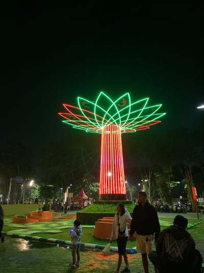 The image depicts a public park scene at night, centered around the Sidoarjo Monument (Monumen Jayandaru) in Sidoarjo, Indonesia. The dominant feature is a tall, stylized tree-like monument. Its cylindrical trunk is vertically striped with red LED lights, while the canopy is formed by layers of neon-green and red light strips arranged in a lotus flower pattern, with stylized shrimp or fish motifs illuminated in gold within the green petals.
In the immediate foreground, a child wearing light-colored clothing stands facing the monument, holding a glowing blue toy. To the child's right, a woman in a white long-sleeved shirt and dark skirt stands holding a white bag. Next to her, a man in a dark hoodie and light shorts stands with a neutral expression. Further right and slightly closer to the viewer, a person in a dark hoodie with "ALAN WALKER" text on the back is crouched or seated on the ground, facing away from the camera.
The ground comprises wet paved walkways and segments of green grass, bordered by a white and blue painted curb. In the mid-ground, green landscaped mounds and orange-colored concrete planters are visible. Various indistinct figures are scattered throughout the park, some walking, others congregated. A red tent structure is present to the left of the monument. Trees line the background, and a large group of motorcycles is parked in the distance. The sky is dark, indicating nighttime conditions.