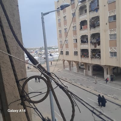 An elevated view in Khan Yunis, Palestinian Territory, captures a street, residential building, and a distant displacement camp under an overcast sky. In the foreground, a textured light-colored wall on the left is partially obscured by multiple dark electrical wires, including a prominent circular coil. "Galaxy A14" is visible in the lower-left corner.
The midground features a paved road with a lighter-colored sidewalk. A tall, grey metal street lamp with a modern fixture stands centrally. To the right, a six-story residential building with a light beige facade, brown vertical strips, and ornate white window frames and balcony railings dominates. Laundry hangs on several balconies. The ground floor is characterized by a series of arched open spaces.
On the street, two individuals dressed in dark abayas walk away from the viewer in the bottom right. Further along the street, a solitary figure in light clothing walks near the building's base. A small group, including a child in pink, walks closer to the building's arched ground floor, and another small group is visible on a dirt path to the left.
The background reveals an extensive area covered with numerous white tents, indicative of a large displacement camp. Beyond the camp, other urban structures and sparse vegetation are visible under a uniformly light grey sky.