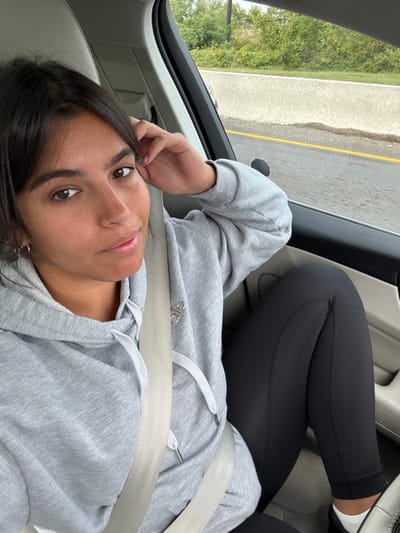 A female individual is seated in the front passenger seat of a vehicle, secured by a light beige seatbelt. She has dark hair and brown eyes, looking directly at the camera with a neutral expression. She is wearing a light grey hooded sweatshirt with an "alo" logo on the left chest, black leggings, and white socks. Her left hand is positioned near her head, with a small silver hoop earring visible in her left ear. The car interior features light-colored trim and upholstery. Outside the passenger window, a highway scene is visible, characterized by an asphalt road with a distinct yellow line, a concrete median barrier, and dense green foliage and trees in the background. This scene is located in West Windsor, United States.
