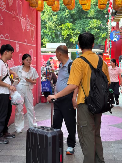 In Yuzhong District, China, a group of people are gathered on a pedestrian walkway. A young woman in white overalls and glasses stands near a young man in a pink t-shirt, both holding mobile phones. Behind them, an older man in a blue shirt faces away from the camera, while another man in a yellow t-shirt with a black backpack also faces away. In the foreground, a black suitcase rests on the ground. The environment features a red wall adorned with heart-shaped drawings and overhead decorative lanterns. Trees and buildings are visible in the background.