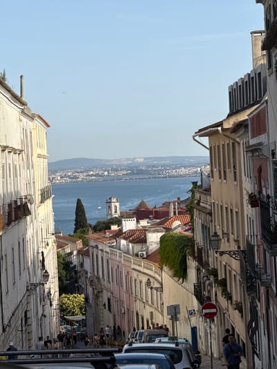 This image depicts a view down a steep, narrow street in Lisbon, Portugal, leading towards a body of water, likely the Tagus River, with a cityscape and hills in the distance. The street is lined with historic buildings, characterized by their colorful facades, balconies, and tiled roofs.
On the left, a tall, cream-colored building with arched windows and ornate balconies dominates the foreground. A satellite dish is visible on one of its balconies. Further down the street, on the right, buildings with beige, pink, and off-white facades are stacked closely together, featuring numerous windows and more balconies. Some buildings have ivy climbing their walls, adding a touch of greenery.
The street itself is paved and appears to be a pedestrian area, with a number of people walking down it. Some are dressed in casual summer clothing. Cars are parked along the sides of the street, including what appears to be a taxi. Traffic signs, including a "no entry" sign and a parking sign, are visible.
In the middle ground, the view opens up to the expansive blue water. A tall, slender evergreen tree stands prominently near the water's edge. A bell tower with a white facade and a clock face rises above the buildings, suggesting a church or cathedral. A white sailboat is visible on the water.
The background shows a sprawling cityscape on the opposite side of the river, with buildings and structures stretching towards the horizon, framed by distant, hazy blue hills. The sky is a clear, bright blue. An airplane is visible as a small dot high in the sky. The overall atmosphere is one of a vibrant, sunny day in a historic European city.