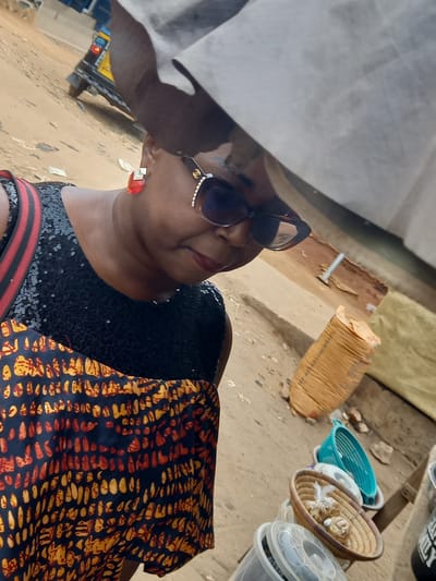 The image presents a close-up, angled portrait of a woman in an outdoor market setting, likely in Onitsha, Nigeria. The woman, of medium complexion, is seen looking slightly upwards and to her right, wearing large, stylish sunglasses with a gold-colored detail on the arm, vibrant red lipstick, and prominent red earrings. She is dressed in a black top or dress heavily adorned with sequins. A richly patterned fabric, featuring orange and yellow abstract designs on a dark background, is draped over her left arm or shoulder, adding a pop of color and cultural flair.
The background depicts a dusty, uneven ground typical of an outdoor market. Various items are scattered on the ground, including a large, light-brown sack, a bright turquoise plastic basin, and a woven straw basket containing small, light-colored objects, possibly shells or crafts. Several clear plastic containers with lids are also visible. In the upper left background, a white tarp or sheet provides shade, beneath which parts of a yellow vehicle, possibly a truck or bus, can be seen. The scene is brightly lit, indicating daytime. The woman appears calm, possibly smiling slightly, suggesting she is either posing or paused in her activity. No explicit text is clearly visible in the image.